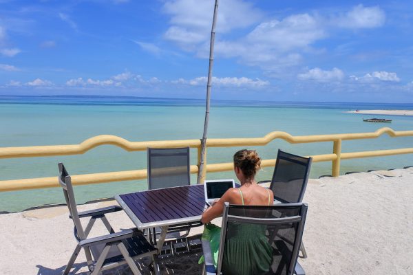 Woman sitting in front of the ocean working on her laptop by the beach.