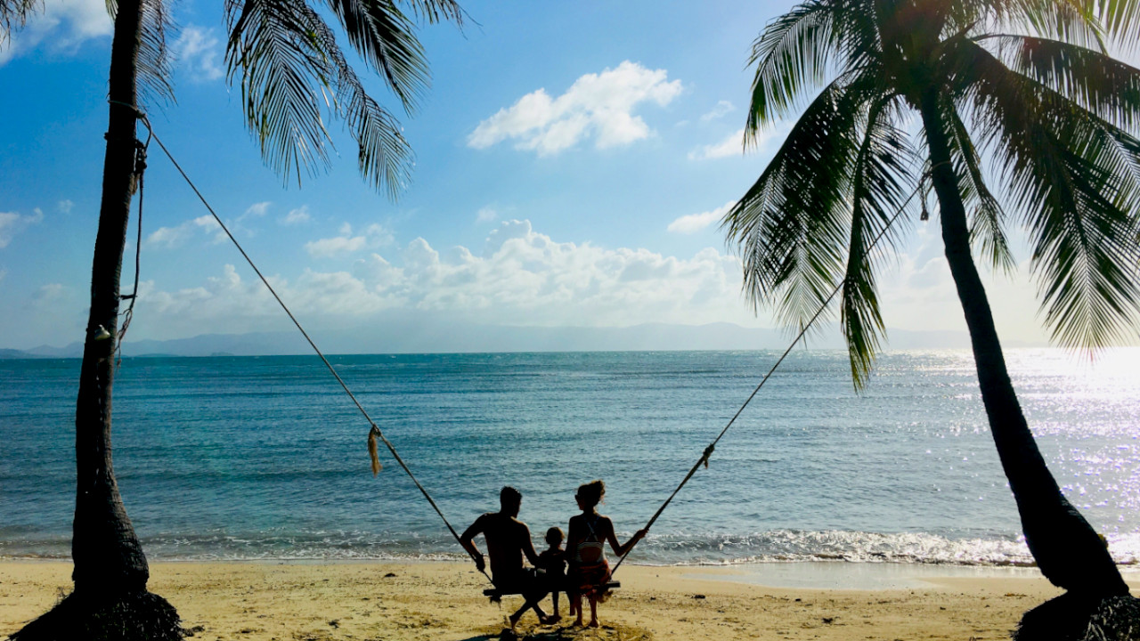Koh Phangan for digital nomad families offer so many cool places like this swing heald by palm trees where a couple with their child sits, the ocean in the background.