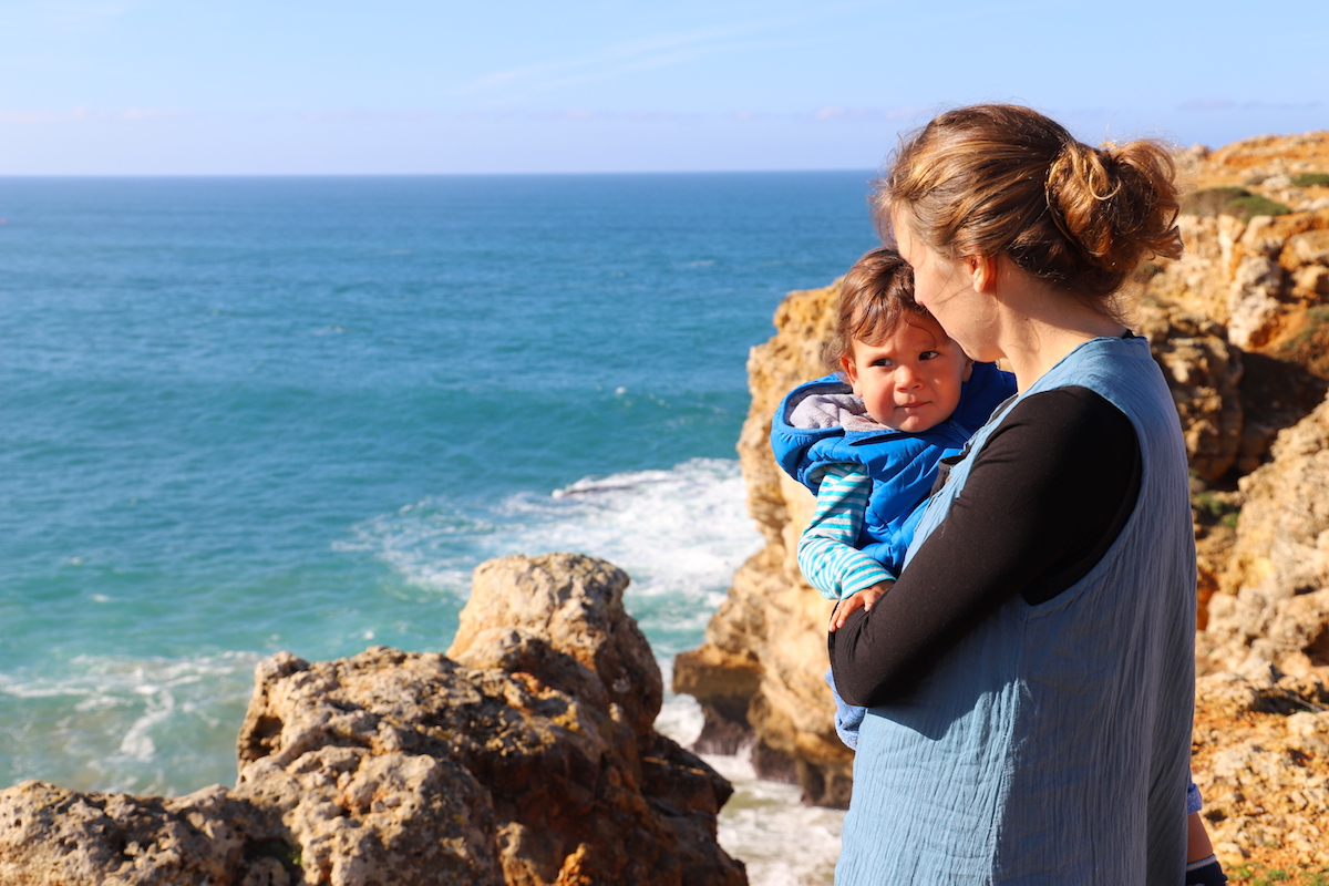 Woman holds a small child in her arms, the ocean and rocks in the background.
