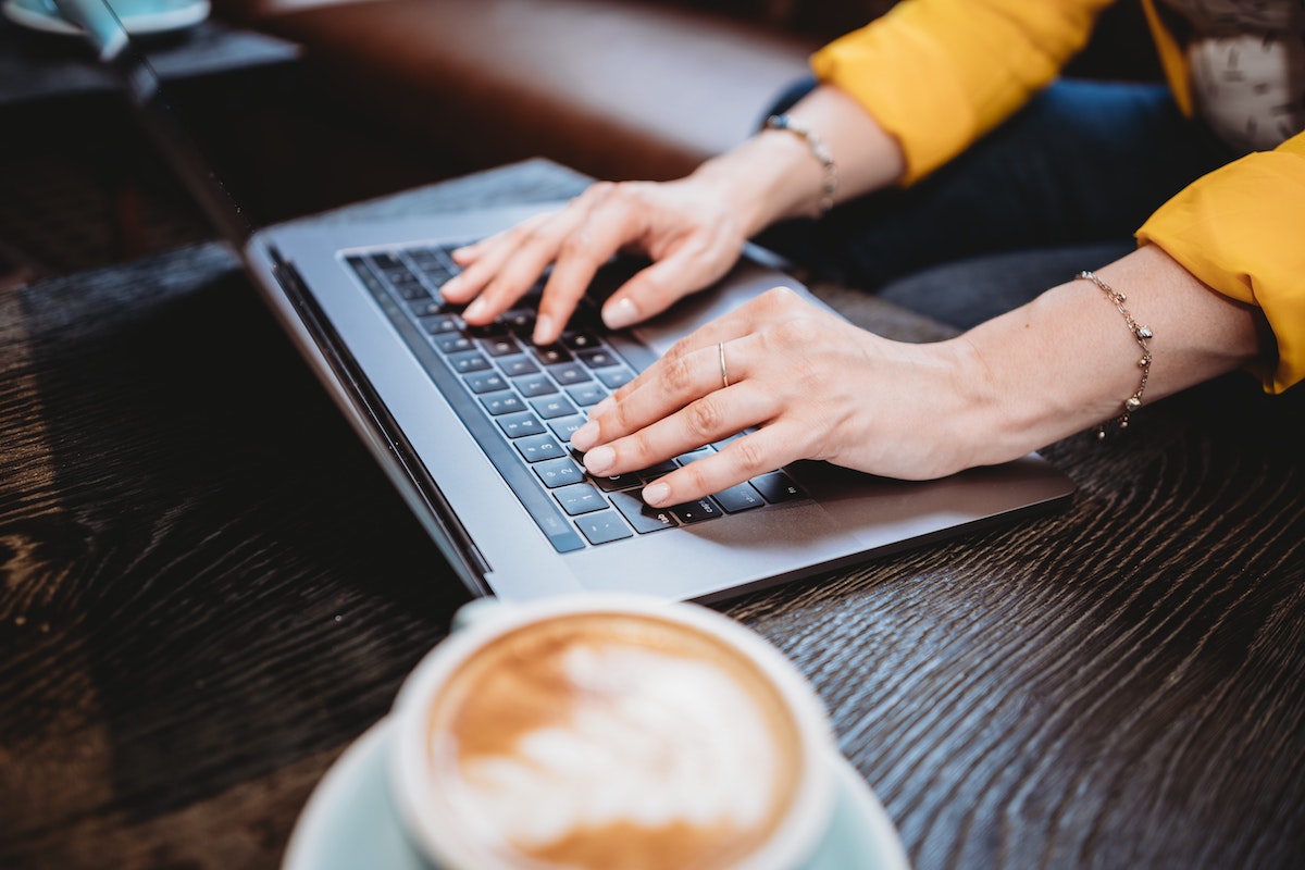 Hands of a woman on opened laptop, a cup of coffee next to her on the table.