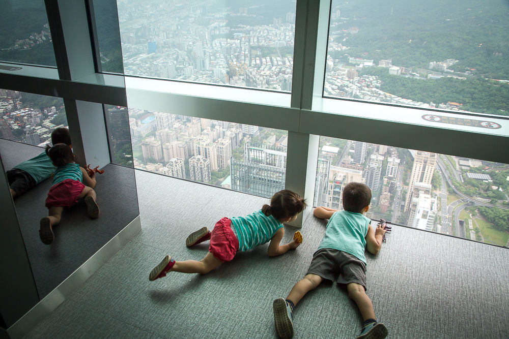 Two young kids laying on the floor of a high building watching outside the window.