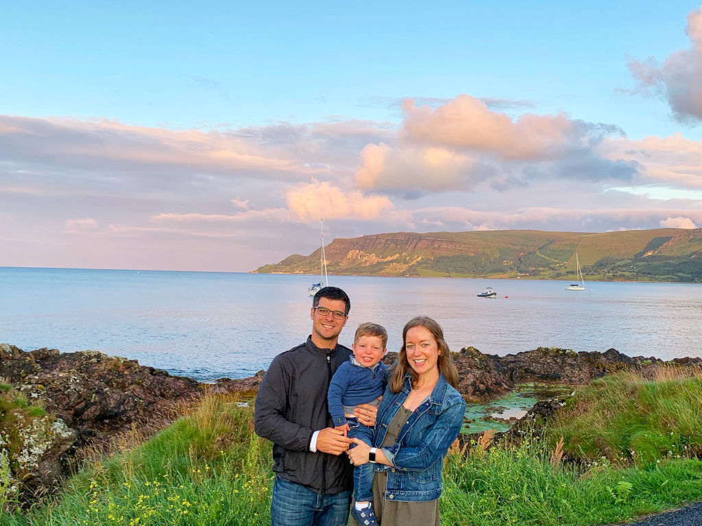 A couple with their toddler standing in front of a scenic landscape, the ocean in the back.