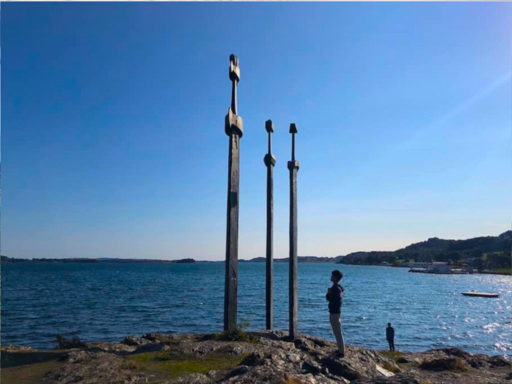 A boy looking up three gigantic swords statues stuck into the ground right by the ocean.