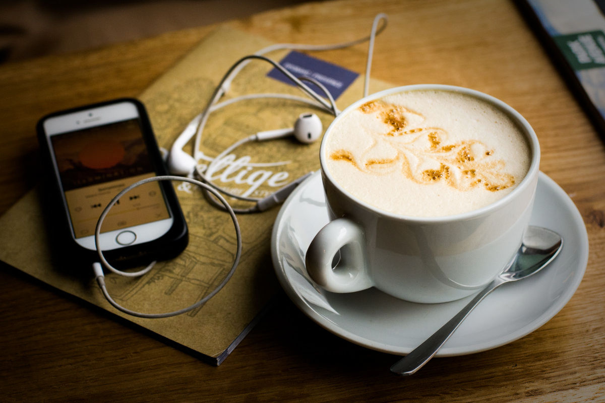 A mobile phone showing a digital nomad podcast on its screen, next to it are headphones, a notebook and a cup of cappuccino on a wooden table. 