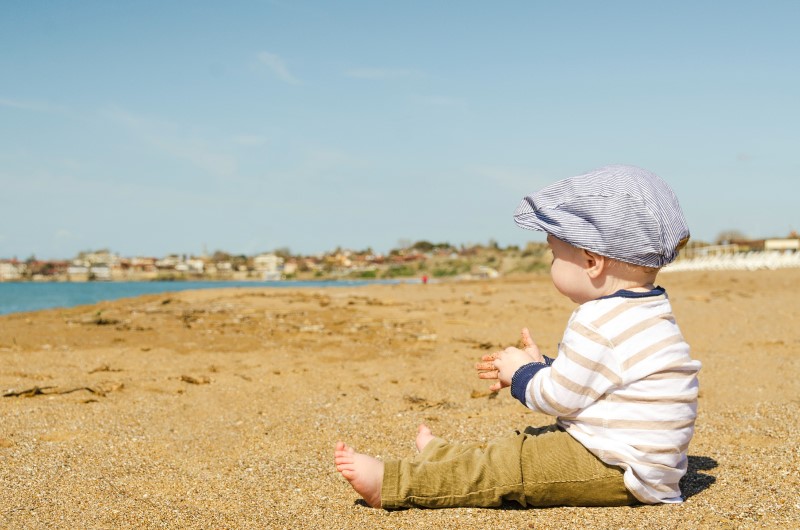 A toddler is sitting at the beach viewing the ocean.